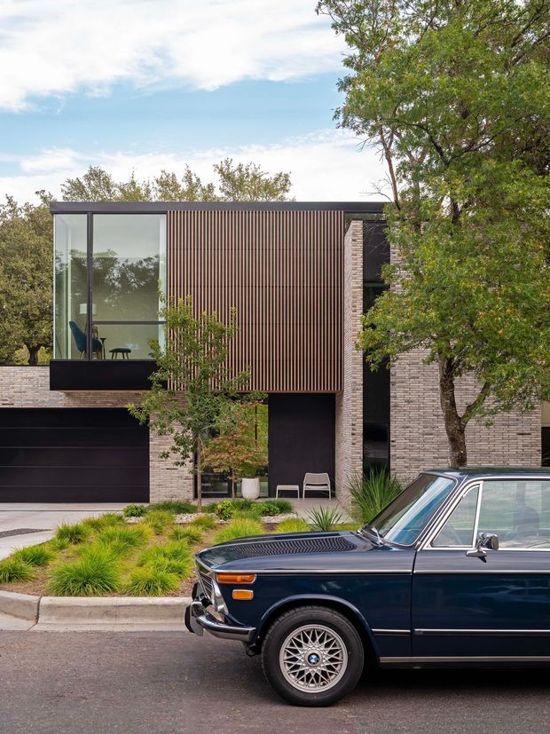 Street-facing view of a refined contemporary multi-level residence with a flat roof, dark horizontal garage door, expansive floor-to-ceiling glazing, vertical timber screening, pale narrow brickwork, and dark metal framing. The landscaped frontage includes ornamental grasses and mature trees. A vintage dark-blue car occupies the foreground street and is not part of the building.