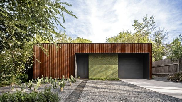 Front exterior view of a low, rectilinear modern structure set within a landscaped residential site. The facade is clad in warm vertical timber battens, with a deeply recessed dark entry or garage zone, a textured green feature wall, and a broad dark garage door. A gravel driveway, concrete path, native planting, cacti, mature trees, and a timber boundary fence frame the building.
