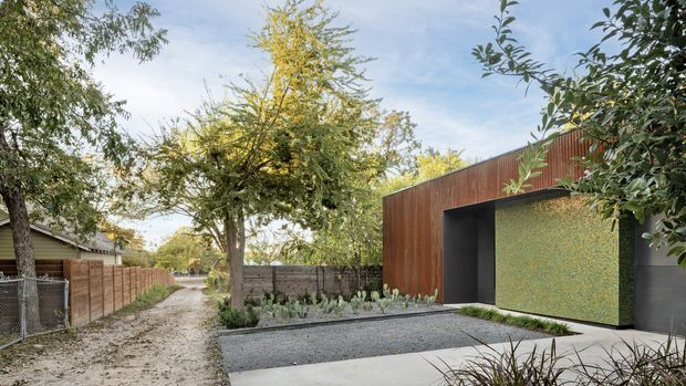 Exterior view of a low, contemporary building set within a landscaped residential garden. The facade combines warm vertical timber cladding with dark recesses and a prominent textured green feature wall, opening onto a pale concrete forecourt and gravel path. Mature trees, planting, and timber boundary fencing surround the structure.