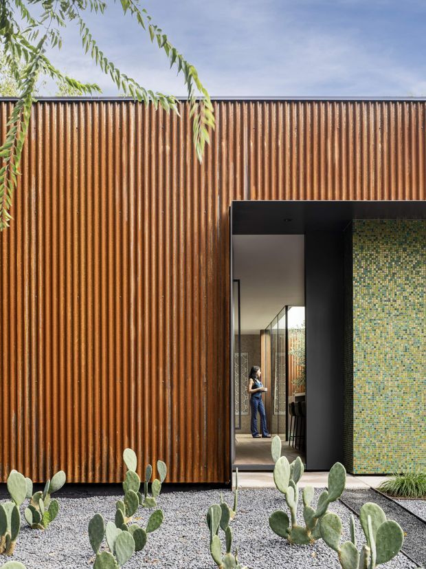 Contemporary architectural exterior featuring a tall rust-toned corrugated metal facade, a deep black-framed entry opening, and a contrasting wall of small green and blue mosaic tiles. The entry reveals a minimalist interior with glazing and a person standing inside. Gravel landscaping, sculptural prickly pear cacti, and overhanging tree branches frame the foreground.