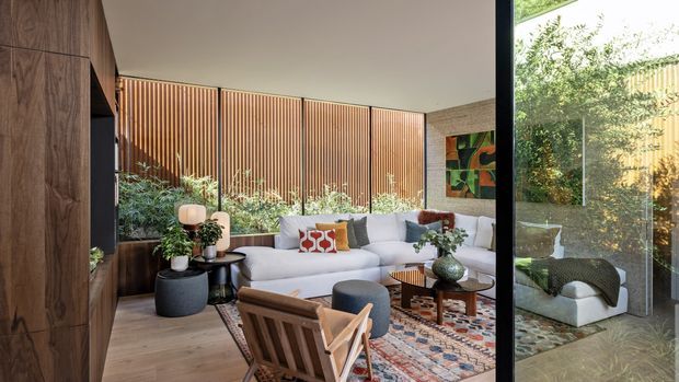 A refined contemporary living room with a large white sectional sofa, lounge chairs, layered patterned rug, coffee tables, indoor plants, and sculptural decor. Warm timber wall panelling and built-in joinery frame the room, while full-height glazing on the right reflects the seating area and opens toward greenery. A vertical timber-slat screen and textured masonry wall add architectural definition.