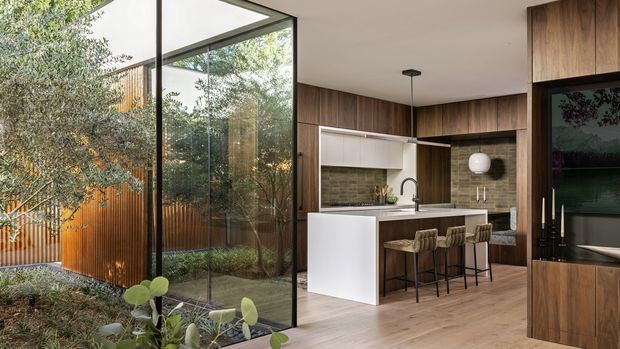 A warm contemporary kitchen and living area with a white island, integrated seating, dark wood cabinetry, and a textured neutral backsplash. Full-height black-framed glazing opens the space visually to a planted courtyard, while built-in timber joinery houses a television on the right. Light timber flooring and restrained pendant lighting create a refined, natural interior.