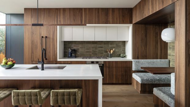 A refined contemporary kitchen with extensive dark timber cabinetry, a large white waterfall island with integrated sink and black tapware, and upholstered island stools. The rear wall features a cooktop, white-framed open shelving, and a textured dark stone or brick-look splashback. To the right, a built-in breakfast nook with grey upholstered seating sits within timber joinery. A linear pendant light and large glazing provide a warm, polished atmosphere.