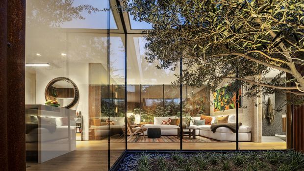 A contemporary living room viewed through expansive floor-to-ceiling glazing, opening onto a landscaped internal courtyard. The space features a low white sectional sofa, warm timber flooring, a sculptural round wall mirror, pale built-in cabinetry, and abundant indoor-outdoor greenery. Reflections in the glazing reveal the room's open, glass-enclosed relationship with the courtyard.