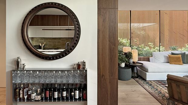 Warm contemporary open-plan interior featuring a built-in home bar beneath a large round dark timber-framed mirror, with a kitchen reflected behind it. A timber-clad structural column separates the bar from a bright living area with a sectional sofa, patterned rug, vertical timber screening, and indoor plants.