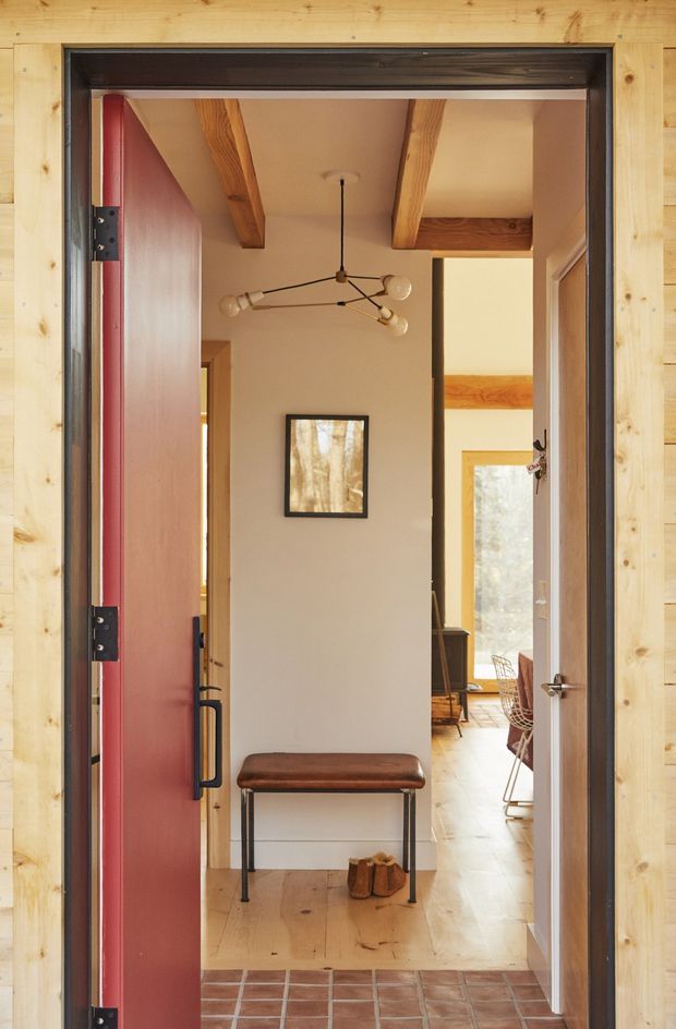 View through an open red entry door into a warm, timber-lined residential entryway. The entry has terracotta square floor tiles at the threshold, pale timber flooring beyond, exposed timber ceiling beams, a slim leather bench, framed artwork, and a contemporary branch-like pendant light. A bright adjoining living or dining area is visible through the interior opening.