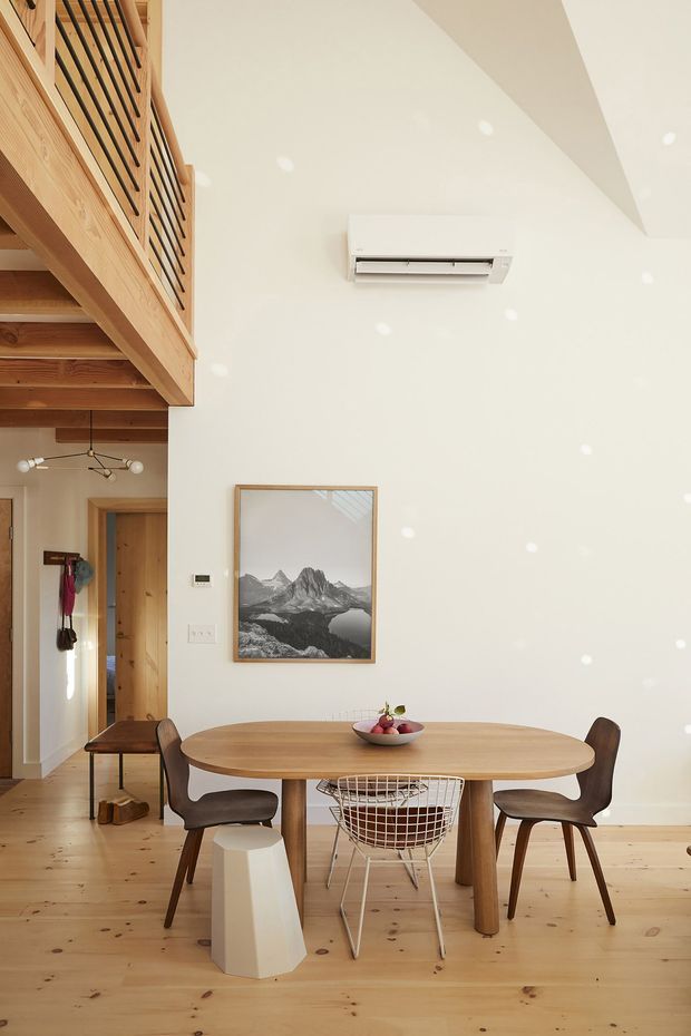 Bright, open-plan dining area with a pale timber floor and a large oval timber dining table surrounded by mixed modern chairs. A timber staircase and mezzanine with dark metal balustrades occupy the left side, while white walls, recessed ceiling lights, a wall-mounted air-conditioning unit, framed mountain artwork, and a doorway complete the space. The room has a warm, minimal contemporary character with strong natural timber detailing.
