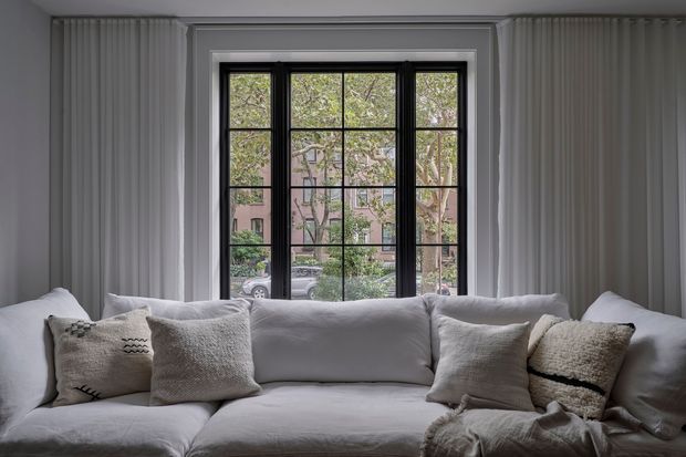 A calm, softly lit living room centered on a large white sectional sofa with textured neutral cushions and a draped throw. Floor-to-ceiling white vertical wall panelling frames a black steel-style multi-pane window, with a leafy residential streetscape visible beyond.