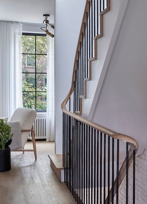 Interior view of a refined stairway beside a tall, black-framed window. The staircase features a warm timber handrail and treads, slender black vertical metal balusters, and white painted walls, with a small upholstered armchair and potted plant nearby. Pale timber flooring, a radiator, and exposed white brick at the lower right add texture. Natural daylight creates a bright, calm atmosphere.