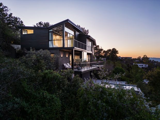 A contemporary multi-level hillside residence photographed at dusk, surrounded by dense vegetation. The dark-clad house features prominent floor-to-ceiling glazing, projecting balconies with slender metal balustrades, and a steeply pitched gable roof over a glazed upper-level living space. Warm interior lighting contrasts with the cool exterior and twilight sky; neighbouring homes and a distant hillside are visible beyond the property.