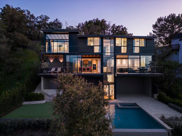 Twilight exterior view of a contemporary multi-level residence set into a landscaped site. The home has a dark rectilinear facade with extensive floor-to-ceiling glazing, multiple balconies and terraces, warm interior lighting, an integrated carport or garage at ground level, and a swimming pool in the foreground.