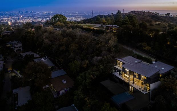 Twilight aerial view of a modern multi-storey residential home set among dense trees on a hillside, with illuminated floor-to-ceiling glazing, a flat roof, and a swimming pool. A sprawling urban cityscape is visible in the distance.
