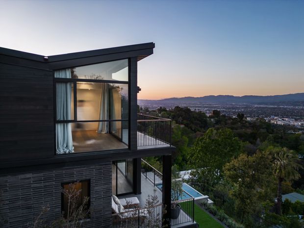 Twilight exterior view of a contemporary multi-level home on a sloped site, featuring dark horizontal cladding, extensive floor-to-ceiling glazing, projecting balconies, and a flat roof. The house overlooks a city basin and distant hills, with a swimming pool and landscaped grounds visible below.