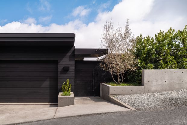 Exterior view of a contemporary dark-clad garage and entry gate, with a flat projecting roofline, concrete planter walls, paved driveway, and minimalist gravel landscaping. The facade is framed by established trees and shrubs beneath a partly cloudy sky.