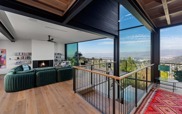 A bright, elevated contemporary living room opening onto a dramatic balcony or terrace with expansive mountain and city views. The room features warm timber flooring and exposed timber ceiling elements, a large deep-green sectional sofa facing a fireplace and built-in shelving, extensive black-framed glazing, and a black metal balustrade around a stair opening or lower-level void. A colourful patterned rug and small side table add accents.