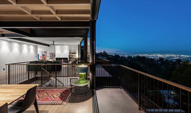 A contemporary split-level residential interior opening onto a spacious balcony or terrace, photographed at dusk. The open-plan kitchen and dining area has white cabinetry, a timber dining table, black chairs, a patterned rug, exposed timber ceiling framing, and dark metal-framed full-height glazing. A metal balustrade borders the terrace, with an illuminated urban landscape visible beyond.