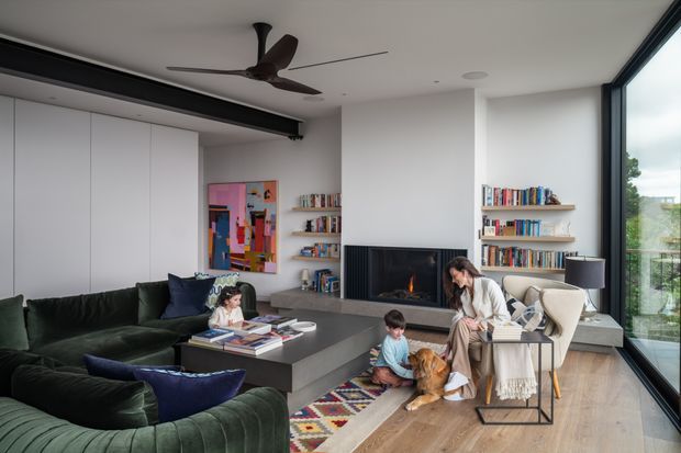 A contemporary open-plan living room with a large dark sectional sofa, low coffee table, built-in white cabinetry, open bookshelves, and a central fireplace. A child and adult are seated near a dog, while floor-to-ceiling glazing opens to an exterior balcony or garden view. The room has timber flooring, a dark ceiling-mounted beam or track element, a ceiling fan, and a colourful area rug.
