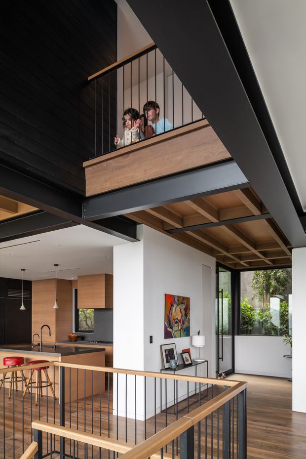 A contemporary double-height interior with an open mezzanine landing overlooking a kitchen and living area. Black steel beams, a black timber-lined ceiling, warm timber flooring and exposed timber ceiling structure contrast with white walls. The mezzanine has a timber fascia and slim black metal balustrade, while the kitchen below features timber cabinetry, dark benchtops and pendant lights. Large glazed doors open toward greenery, and framed artwork and console furniture add detail.