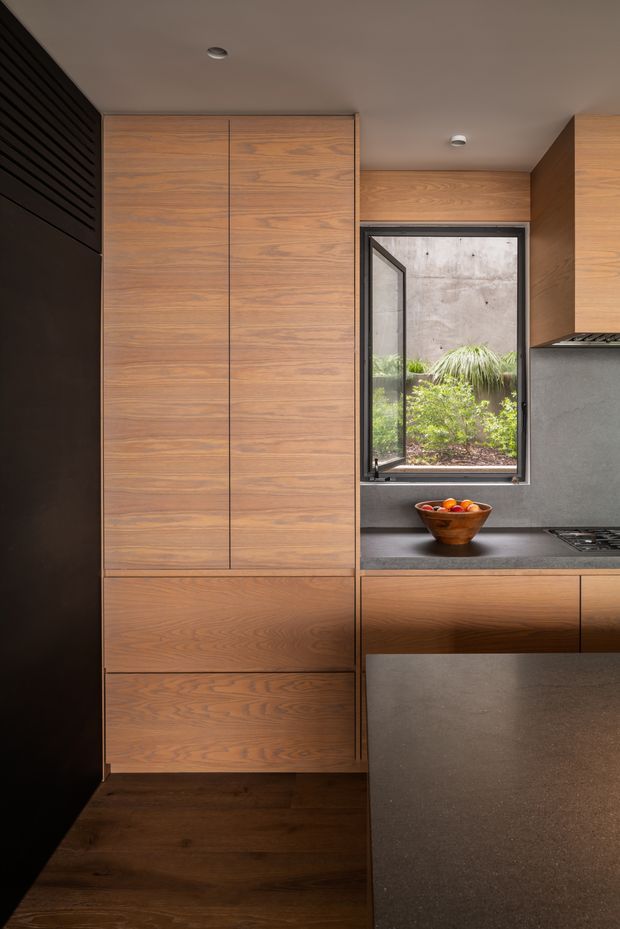 A compact contemporary kitchen with full-height warm timber cabinetry, dark stone benchtops, integrated appliances, and a black-framed window overlooking a planted courtyard. The space has a restrained, refined material palette with recessed ceiling downlights and dark timber flooring.
