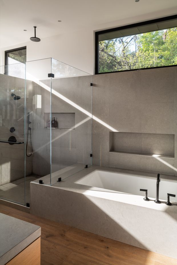 Contemporary bathroom featuring a large integrated bathtub and separate frameless glass shower enclosure. Warm grey stone-look wall and bath surfaces, recessed niches, matte black tapware and shower fittings, a black-framed horizontal window, and timber flooring create a refined minimalist interior. Strong diagonal sunlight enters through the glazing and illuminates the room.
