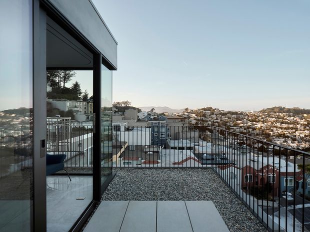A contemporary rooftop terrace viewed from the edge of a glazed building. The terrace has a gravel-covered surface with large rectangular concrete pavers, slim black metal balustrades, and full-height dark-framed glazing opening to the interior. A dense hilly urban neighbourhood is visible beyond.