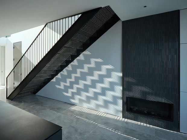 A modern interior stairway with a slim black metal balustrade and perforated steel stair stringer casting geometric shadows across a pale wall. The stair rises beside a dark vertically ribbed feature wall containing a built-in linear fireplace. Large-format grey flooring and clean white surfaces create a minimalist, high-contrast composition.
