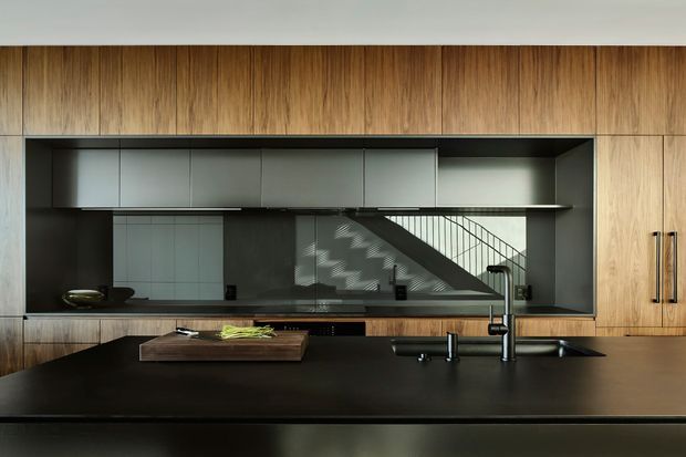 A sleek contemporary kitchen with a large dark island bench in the foreground, integrated black sink and tapware, timber veneer cabinetry and wall panels, and a dark framed splashback opening. Grey overhead cabinetry and a reflective glazed surface reveal a staircase beyond, adding depth and architectural interest. The restrained palette and clean lines create a refined, minimalist atmosphere.