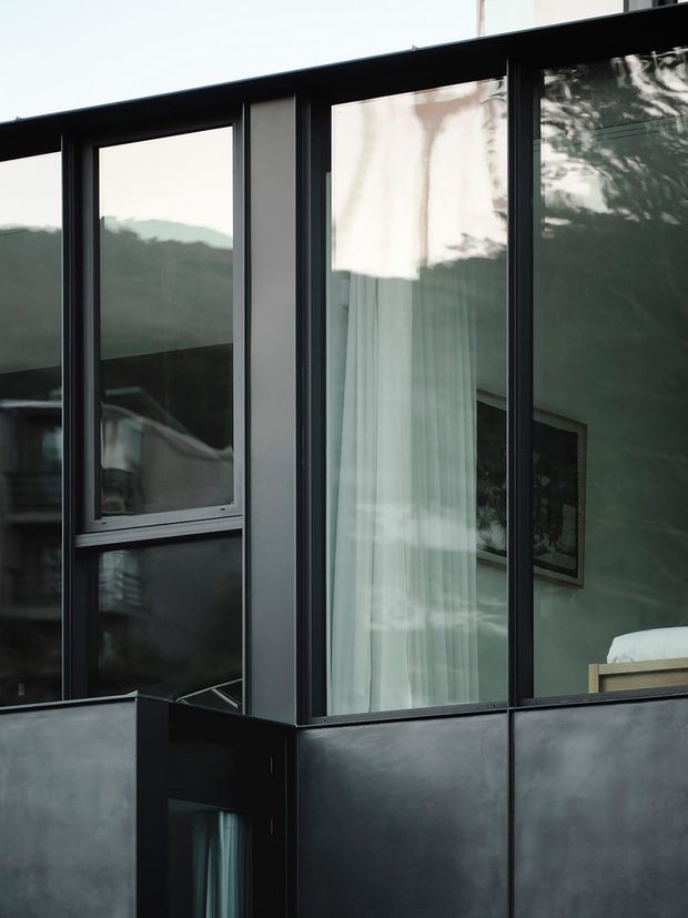 Close-up exterior view of a contemporary building corner with dark metal-framed floor-to-ceiling glazing and solid dark cladding panels. Curtains, interior artwork, and part of a bed are visible through the windows, while reflections show surrounding trees and hills. The composition emphasizes the facade, glazing, and material contrast.