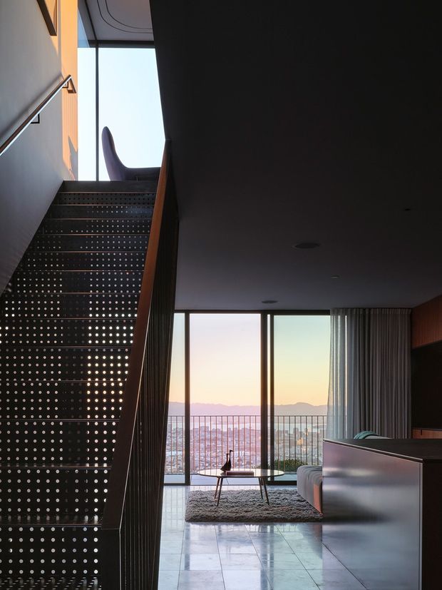 A dramatic interior view looking up a perforated metal staircase toward a bright upper-level window, with a living area beyond. Floor-to-ceiling glazing opens to a balcony and distant mountain-and-city views. The living room contains a low coffee table, rug, sofa, and dark built-in joinery, creating a refined, moody contemporary atmosphere.