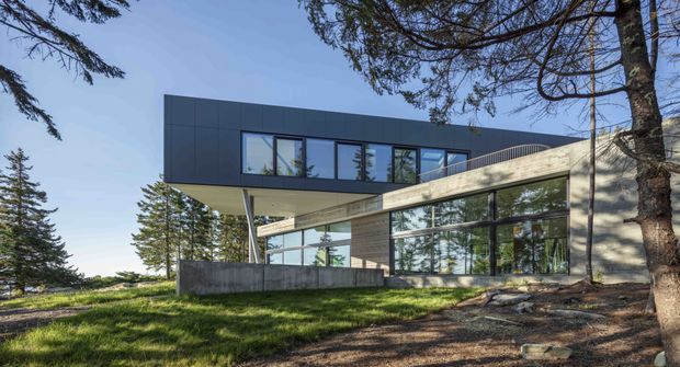 Exterior view of a contemporary two-storey home set among mature trees, featuring a dark charcoal upper volume, extensive floor-to-ceiling glazing, a projecting angled concrete or light-toned masonry element, and a landscaped sloping site.