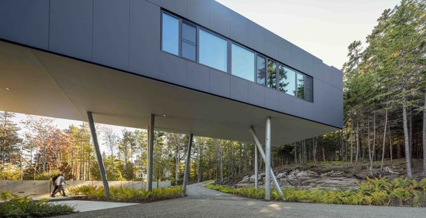 Exterior architectural view of a contemporary dark-clad elevated building set within a wooded, rocky landscape. The upper rectangular volume projects outward over a covered ground-level walkway or entry area, supported by slender angled metal columns. A horizontal band of large windows reflects the surrounding trees, while a paved path leads beneath the cantilevered form.
