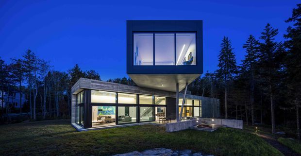 A modern two-storey house at dusk, set within a wooded landscape. The lower level is extensively glazed with warm interior lighting, while a dark rectangular upper volume projects over the terrace on slender supports. The flat-roofed composition, concrete terrace edge, expansive windows, and surrounding trees create a dramatic contemporary architectural exterior.