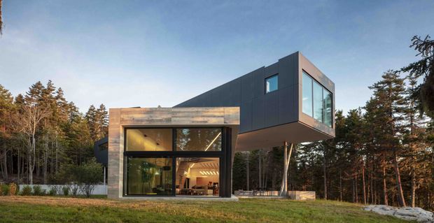 A modern two-storey residence set within a wooded landscape. The building combines a dark charcoal-clad upper volume with a projecting glazed corner supported by slender columns, over a lower section featuring warm timber or wood-toned cladding and expansive floor-to-ceiling glazing. Interior lighting and furnishings are visible through the glass, while the surrounding forest and low roofline emphasize a contemporary, site-responsive design.