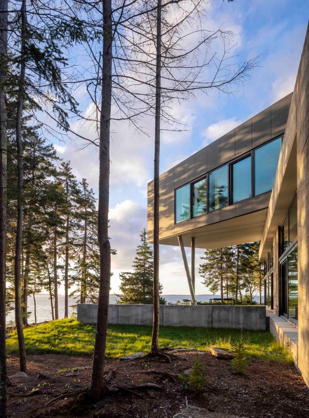 Exterior architectural photograph of a contemporary box-like house set among tall trees beside a visible body of water. The building features dark metal cladding, extensive blue-tinted glazing, a projecting upper volume supported by slender angled columns, and a concrete retaining or boundary wall. A covered outdoor circulation area and glazed ground-floor facade are visible on the right.