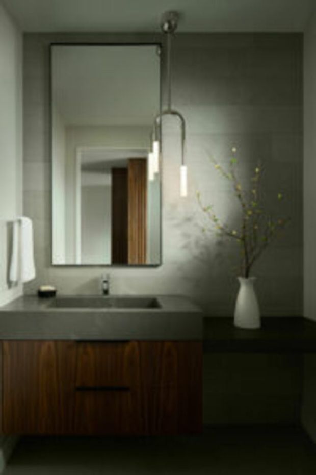 A moody, minimalist bathroom vanity with a large frameless rectangular mirror, grey stone or concrete-look benchtop and integrated basin, dark grey wall finish, and warm dark timber cabinetry below. A simple pendant light and white ceramic vase provide subtle contrast.