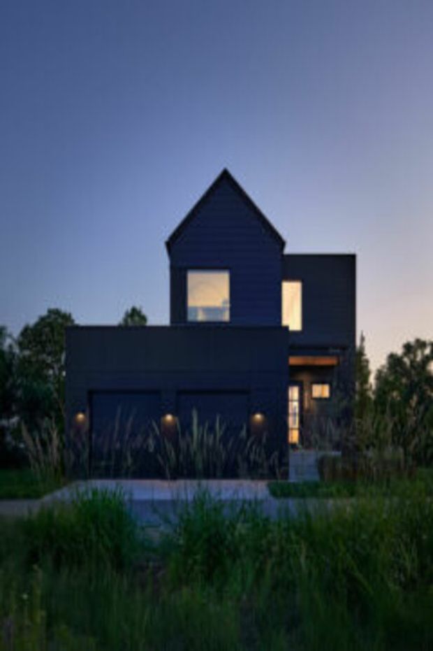 A dusk exterior view of a contemporary multi-storey house with a dark, boxy facade and a prominent steep gabled volume. Warm light glows from the windows and recessed entry, while tall ornamental grasses and a simple paved path frame the foreground.