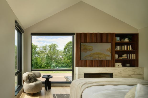 A bright contemporary bedroom with a high sloped ceiling, expansive black-framed glazing, and views of greenery. The room features a low upholstered bed in light neutral bedding, a sculptural lounge chair beside a small dark side table, pale timber flooring, and a built-in feature wall combining vertical timber battens, open shelving, artwork, and a marble-clad fireplace surround.