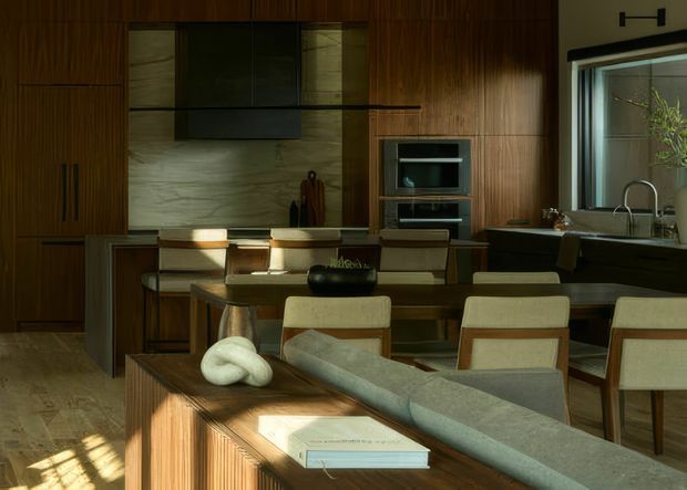 A warm, moody open-plan kitchen and dining area with extensive dark timber wall panelling and cabinetry. The kitchen includes a central island with integrated seating, a dark rangehood and stone-look splashback, built-in appliances, and a sink beneath a large window. A timber dining table with upholstered chairs occupies the foreground, with a sculptural sofa and built-in timber joinery creating separation from the dining zone. Sunlight creates strong highlights and shadows across the timber surfaces and floor.