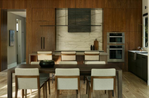 A refined open-plan kitchen and dining area with a long dark timber dining table and upholstered dining chairs in the foreground. The kitchen is integrated into a full-height warm timber wall with vertical fluted detailing, concealed cabinetry, black handles, and built-in stainless-steel appliances. A large veined light stone backsplash and dark central rangehood create a strong feature wall, while pale timber flooring and restrained contemporary furnishings complete the space.