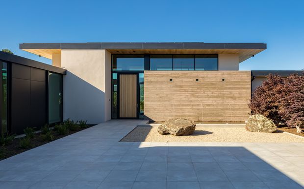 Modern single-storey house facade viewed from a broad paved front entry court. The building has a strong horizontal flat roof with deep eaves, dark framed glazing, a tall timber front door, smooth light rendered walls, and a prominent horizontal timber-clad feature wall. Gravel landscaping with boulders and sculptural planting adds a restrained contemporary character.