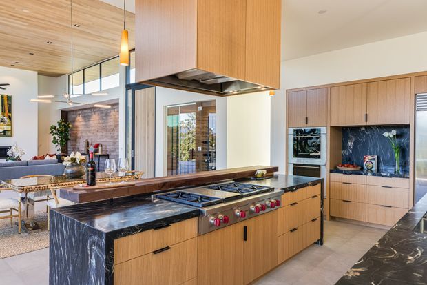 Bright open-plan kitchen with a large timber island, dark veined stone waterfall ends and benchtops, integrated professional range cooker, and a suspended timber rangehood enclosure. Floor-to-ceiling timber cabinetry continues along the rear wall, with a dark blue-grey stone splashback and integrated appliances. The kitchen opens to a contemporary living area with timber ceiling lining, glazing, and pendant lighting.