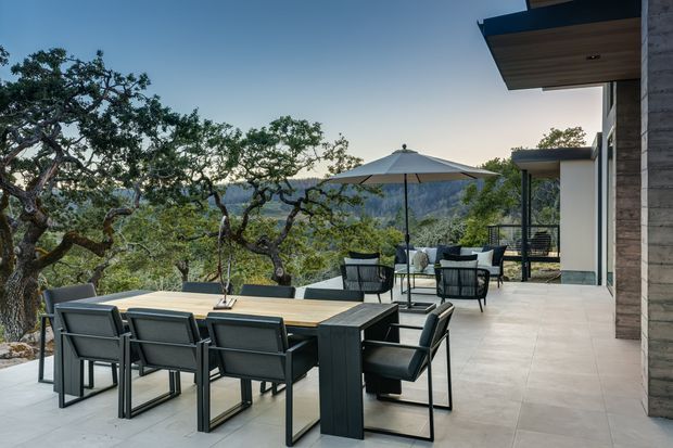 An expansive outdoor terrace overlooking a wooded mountainous landscape, furnished with a large rectangular dining table and chairs in the foreground, plus a smaller lounge setting beneath a central umbrella. The contemporary building has broad roof overhangs, light rendered walls, dark-framed glazing, and a textured timber-clad column. Large-format pale tiled paving unifies the terrace, creating a refined indoor-outdoor entertaining space.