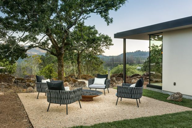 A contemporary outdoor seating area beside a minimalist single-storey house, set on a landscaped gravel terrace among mature trees and rocky terrain. The space includes four woven lounge chairs arranged around a low circular fire bowl, with expansive glazing and a dark projecting roof edge on the building. A wooded, hilly landscape is visible beyond.