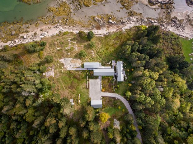 Aerial view of a contemporary building complex set within dense vegetation beside a rocky shoreline and shallow green water. The property includes several connected rectilinear structures with pale roofs, a broad paved approach, and a curved driveway through the surrounding landscape.