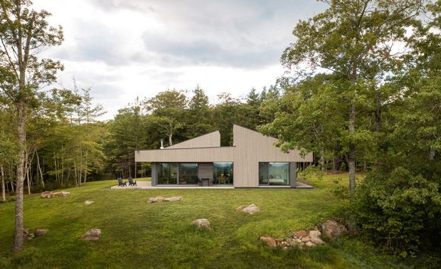 A low-profile contemporary home set within a wooded landscape, viewed from the lawn. The facade combines broad floor-to-ceiling glazing with vertically ribbed light-coloured cladding and a sharply asymmetric pitched roof form. Large areas of lawn, scattered natural stones, and mature deciduous trees surround the building.