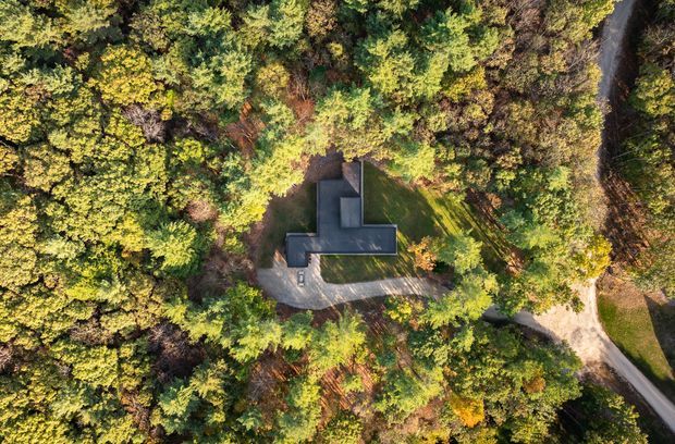 Aerial view of a dark-roofed, cross-shaped building nestled within dense mature woodland. Curving driveways or paths approach the structure from the right and lower edge, while the surrounding tree canopy largely conceals the site.