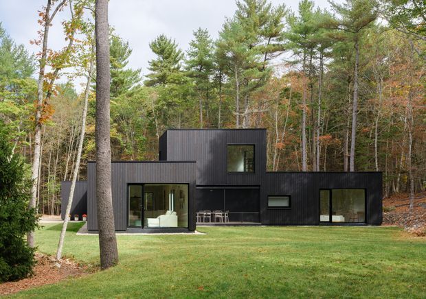 A modern, single-storey dark-clad house set within a wooded landscape. The building is composed of simple interlocking rectangular volumes with flat roofs, large dark-framed glazed openings, and a sheltered outdoor dining area. A broad lawn leads up to the house, which is surrounded by mature pine and deciduous trees.