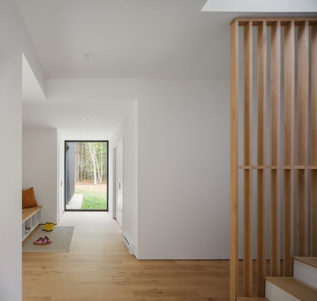 A bright, minimalist residential entry hall with pale timber flooring and white walls. The hallway leads to a full-height glazed door opening toward a wooded outdoor area. A built-in bench with a cushion and shoe storage sits to the left, while a tall vertical timber slat screen partially conceals the stairway on the right. The space has clean lines, recessed ceiling lights, and a light neutral palette.