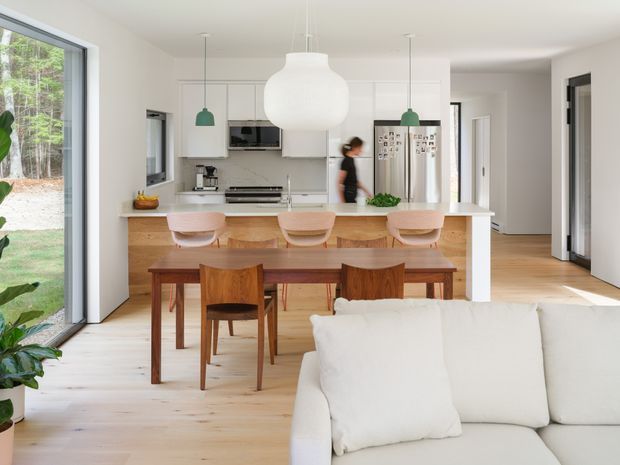 Bright open-plan kitchen and dining area viewed from the living room. The space features white flat-panel cabinetry, a pale benchtop and splashback, a timber island with three light-coloured stools, a timber dining table and chairs, pale timber flooring, large windows and glazed doors, and simple pendant lighting. A white sofa occupies the foreground, while a hallway is visible beyond the kitchen.