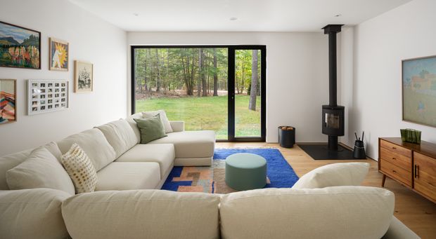 A bright, contemporary living room with a large cream sectional sofa arranged around a round ottoman and colourful area rug. Black-framed floor-to-ceiling glazing opens to a wooded garden, while a freestanding black wood-burning stove, timber flooring, white walls, framed artwork, and a mid-century-style timber sideboard define the room.